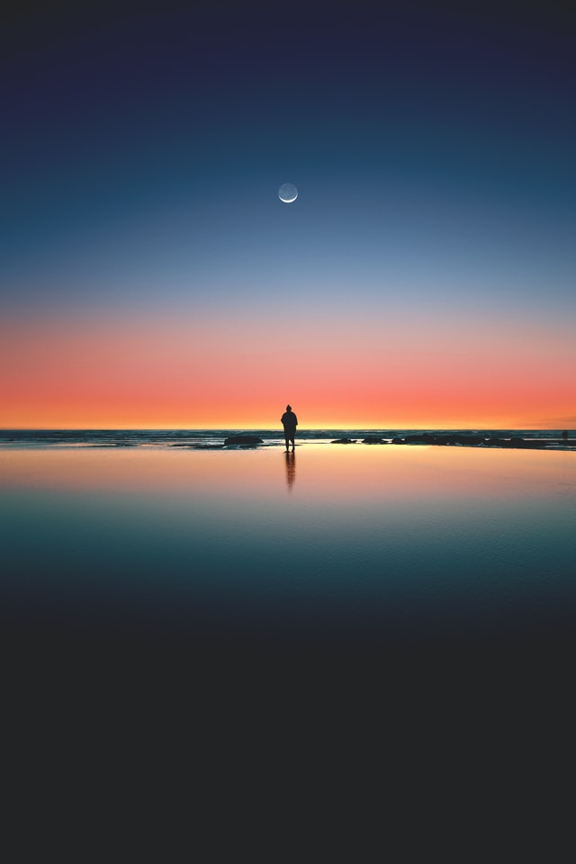 A silhouette of a person looking out at a sunset under the moon