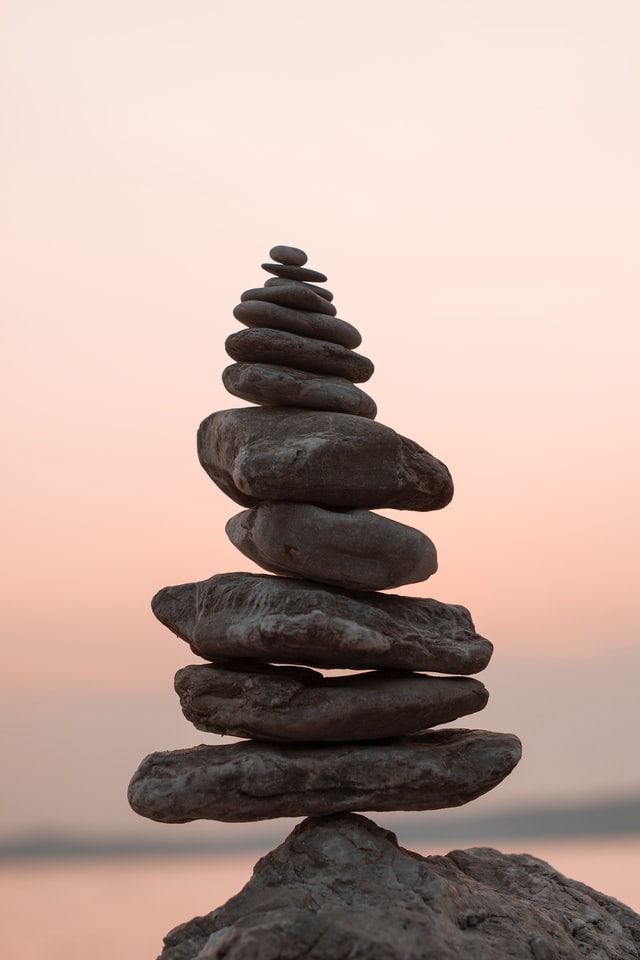 A pile of flat rocks balancing on a peak point of a larger rock with a blurred beach in the background